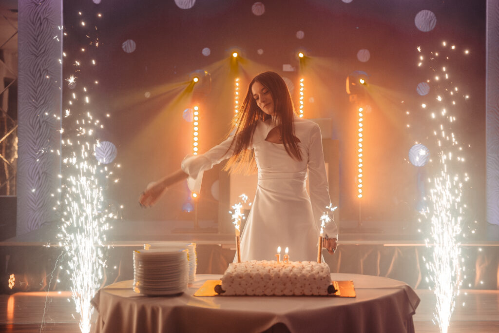 Brunetka w białej sukni stojąca przy torcie urodzinowym, za nią fajerwerki