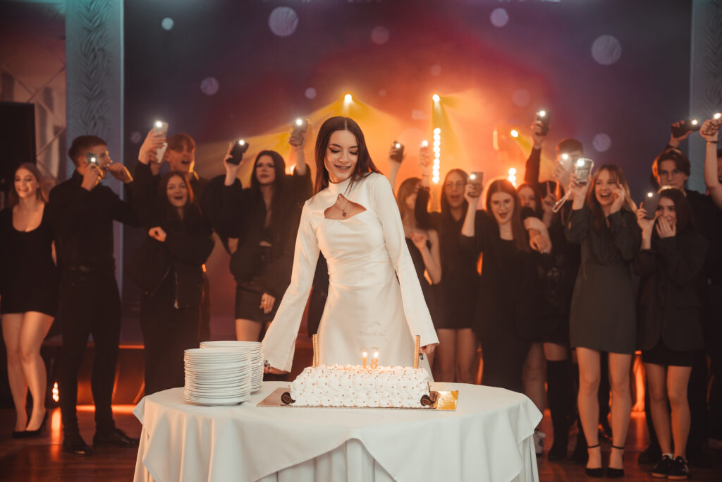 Brunetka stojąca przy torcie urodzinowym, w tle tłum ludzi podczas zabawy