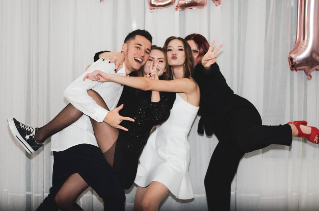 Czwórka młodych ludzi pozuje radośnie do zdjęcia na imprezie – dwie kobiety i dwóch mężczyzn, wszyscy uśmiechnięci i przytuleni, w energicznej, tanecznej pozie. Tło stanowią białe zasłony i dekoracje balonowe.