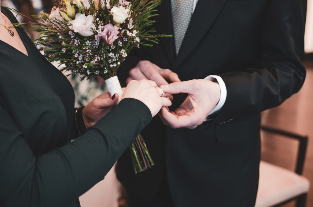 Para podczas ceremonii ślubnej – mężczyzna w garniturze zakłada kobiecie pierścionek na palec. Kobieta trzyma bukiet kwiatów z różowymi i białymi akcentami.