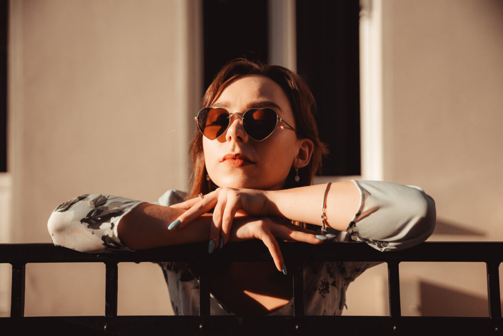 Młoda kobieta w okularach przeciwsłonecznych opierająca się o barierkę balkonu, korzystająca ze słońca i pięknej pogody na balkonie
