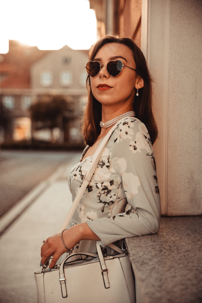 Młoda kobieta pozująca na tle miejskiej scenerii w słoneczny dzień, ubrana w elegancką sukienkę i okulary w kształcie serca, trzymająca białą torebkę.