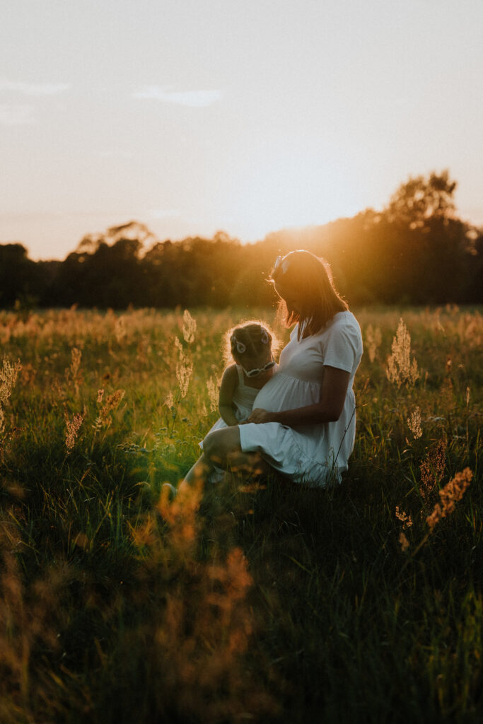 Kobieta w ciąży siedząca na łące z dzieckiem, uchwycona w ciepłych promieniach zachodzącego słońca podczas sesji zdjęciowej.