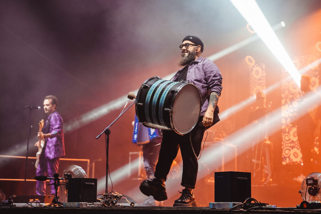 "Muzyk w fioletowej koszuli grający na dużym bębnie podczas koncertu. Na scenie widać intensywne światła i dym sceniczny, a w tle kolejnych członków zespołu.