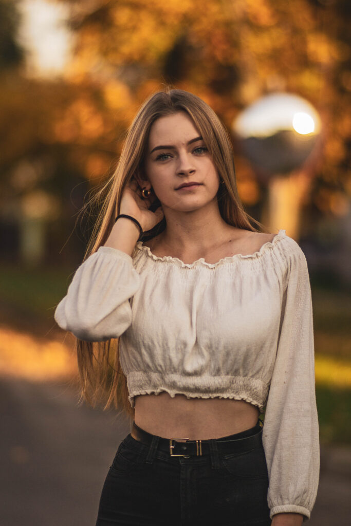 Młoda kobieta z długimi, prostymi włosami pozuje na zewnątrz przy ciepłym, jesiennym świetle. Ma na sobie jasną, długorękawną bluzkę odsłaniającą brzuch i delikatny, spokojny wyraz twarzy.