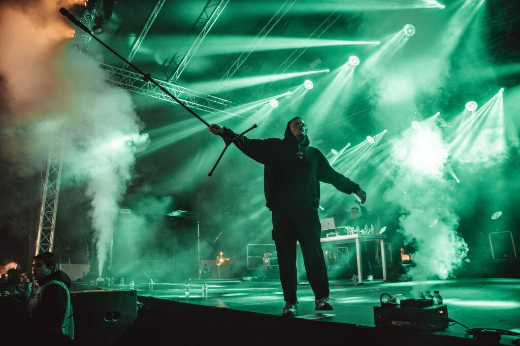 Artysta stojący na scenie podczas koncertu, wystawiający stelaż z mikrofonem w stronę publiczności. W tle widać dużo mgły i dymu scenicznego, przeważające zielone kolory.