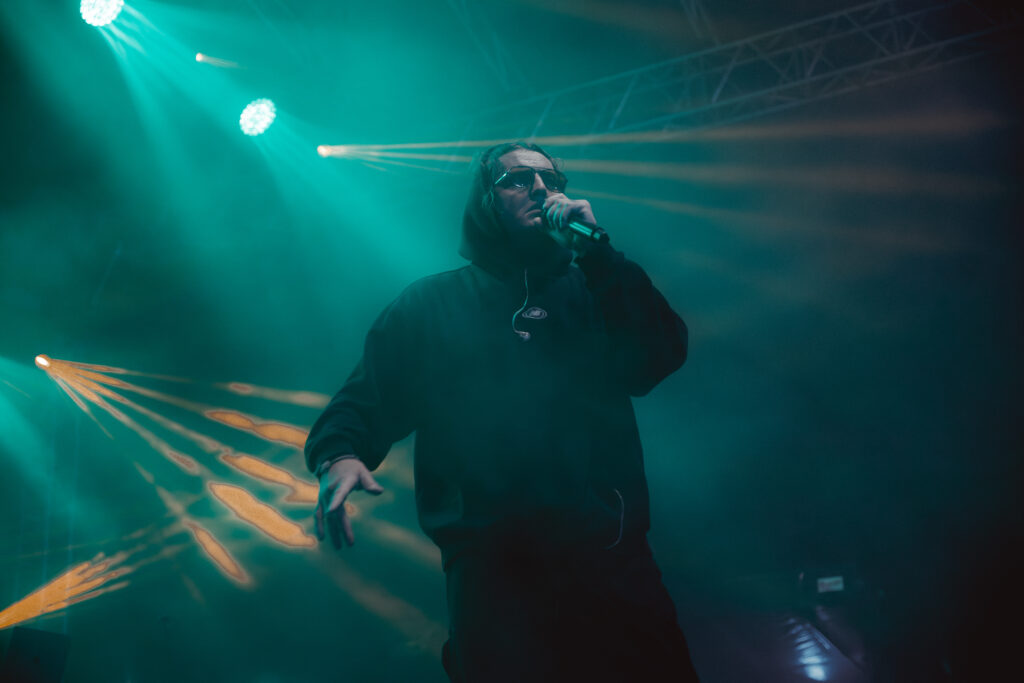 Raper ubrany na czarno, z kapturem na głowie i okularami przeciwsłonecznymi, występuje na scenie podczas koncertu. Trzyma mikrofon i śpiewa, a za nim widać intensywne, kolorowe światła sceniczne w odcieniach niebieskim i pomarańczowym oraz lekką mgłę sceniczną.