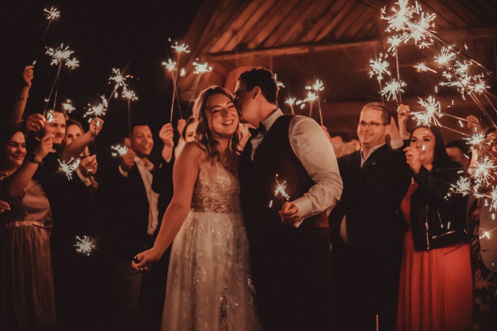 Młoda para podczas wesela, otoczona gośćmi trzymającymi zimne ognie, celebrująca moment miłości i radości.