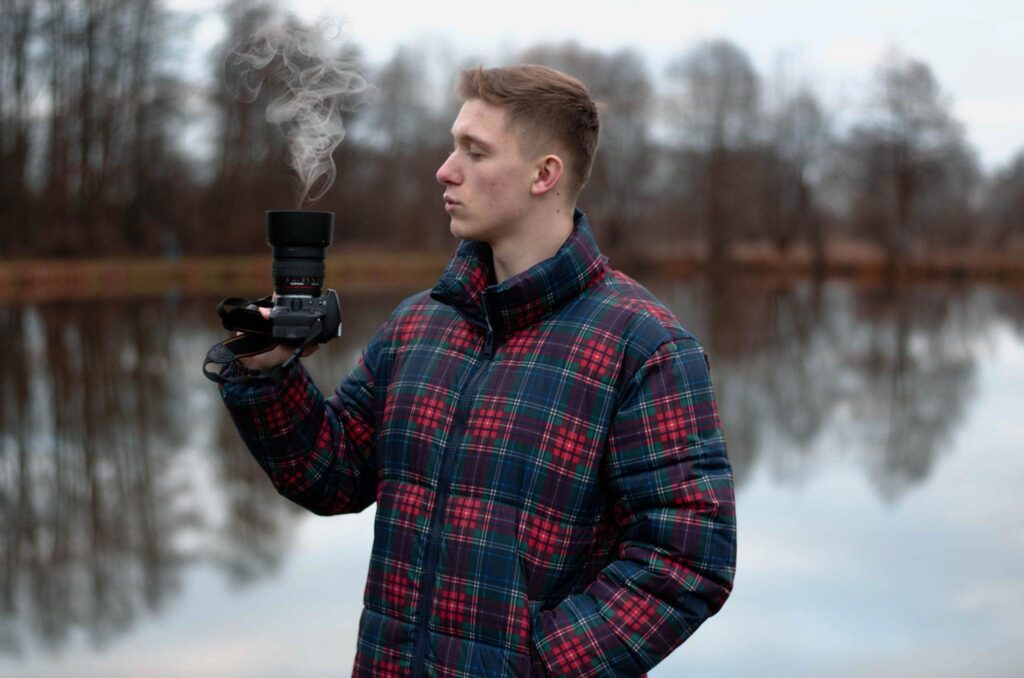 Mężczyzna w kraciastej kurtce trzyma aparat, z którego unosi się dym, w tle jezioro i drzewa.